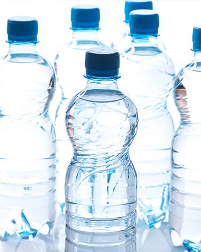 Six clear plastic water bottles with blue caps are arranged close together. The bottles are filled with water and placed on a white surface with a white background.