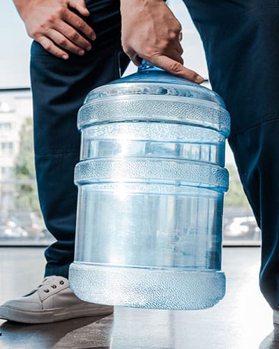 A person wearing dark pants and white sneakers is holding and carrying a large, transparent blue water jug by the handle indoors.